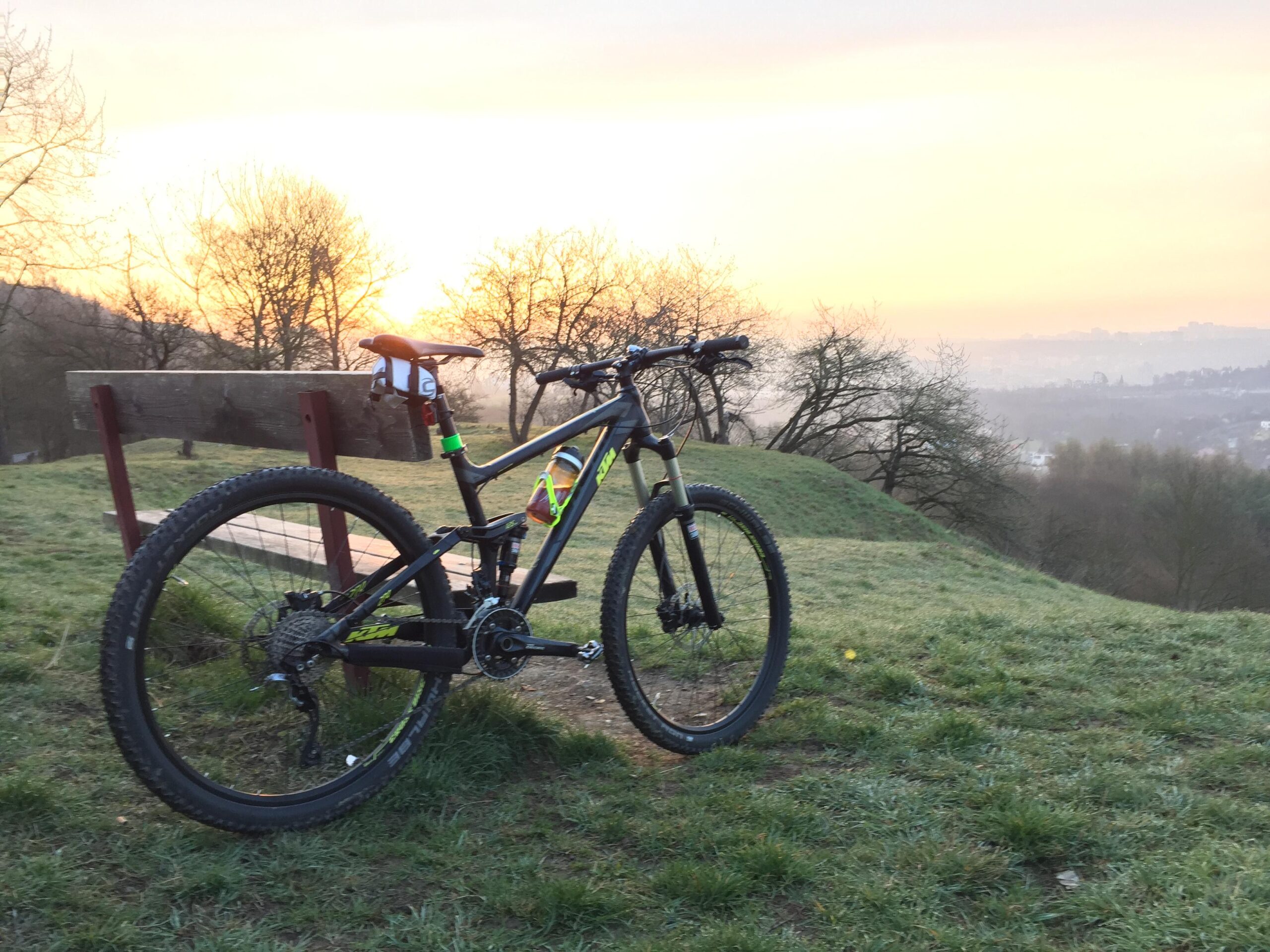 Ktm Lycan 274: A mountain bike parked on a grassy hillside with a wooden bench in the background, set against a glowing sunrise. The scene features silhouettes of trees and a misty landscape in the distance, creating a serene and inviting outdoor atmosphere.
