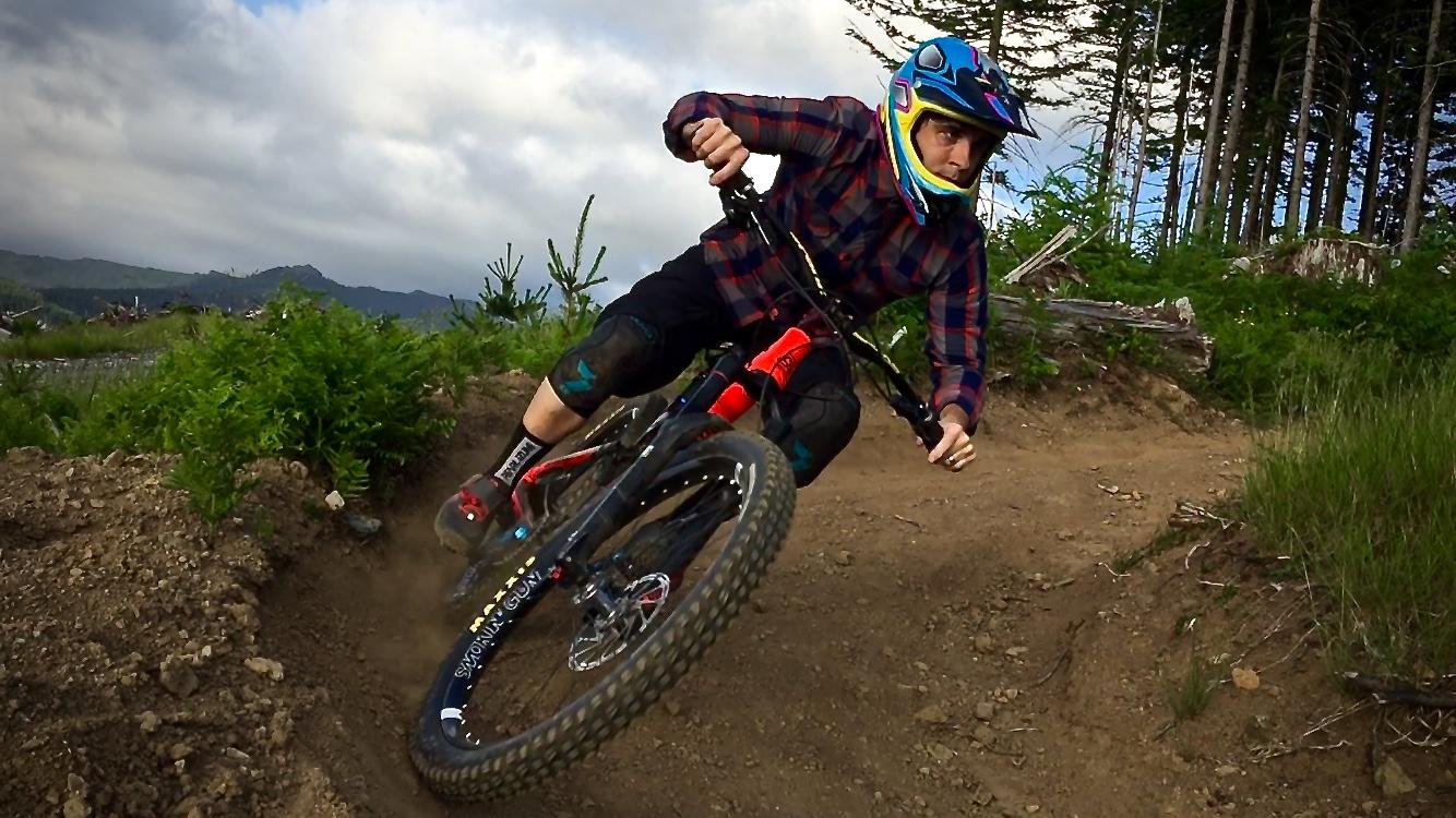 A mountain biker in a plaid shirt and helmet leans into a tight turn on a dirt trail in a lush green forest. The rider is positioned low, with one knee close to the ground, as the bike tires kick up dust, showcasing the intensity of the ride. Pine trees and mountains are visible in the background under a cloudy sky. Cold Creek mountain bike trail.