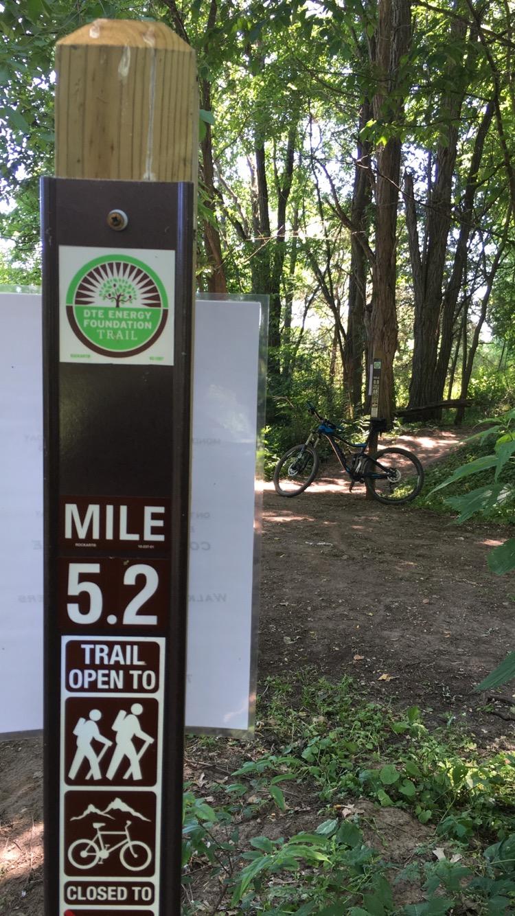 A trail marker indicates Mile 5.2 on the DTE Energy Foundation Trail, with instructions showing the trail is open to hikers and closed to cyclists. In the background, a blue bicycle is parked beside the trail entrance, surrounded by trees and greenery. DTE Energy Foundation Trail mountain bike trail.