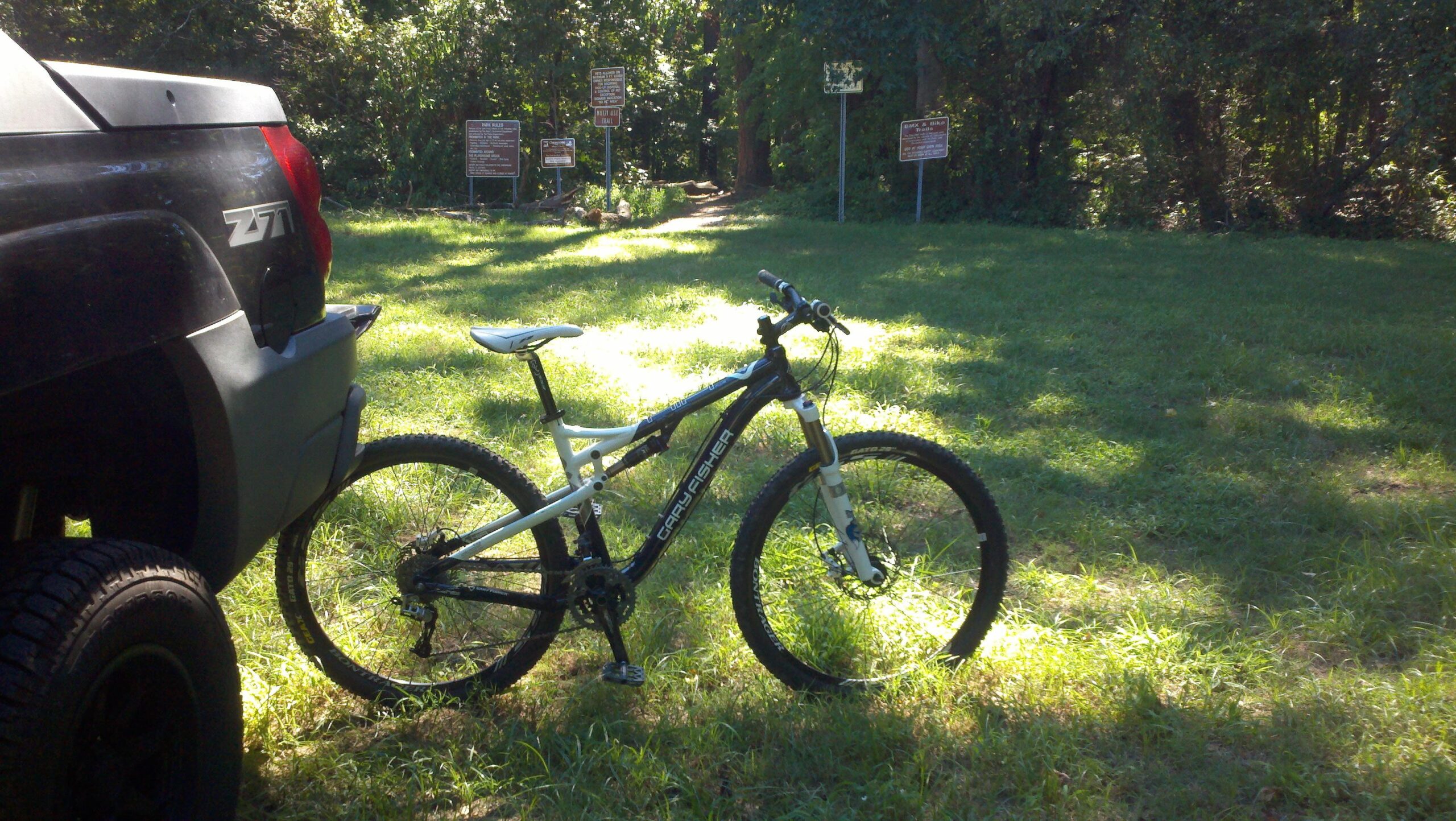 A mountain bike parked on grass next to a black vehicle, with wooded paths and multiple informational signs visible in the background. Indian River Park mountain bike trail.