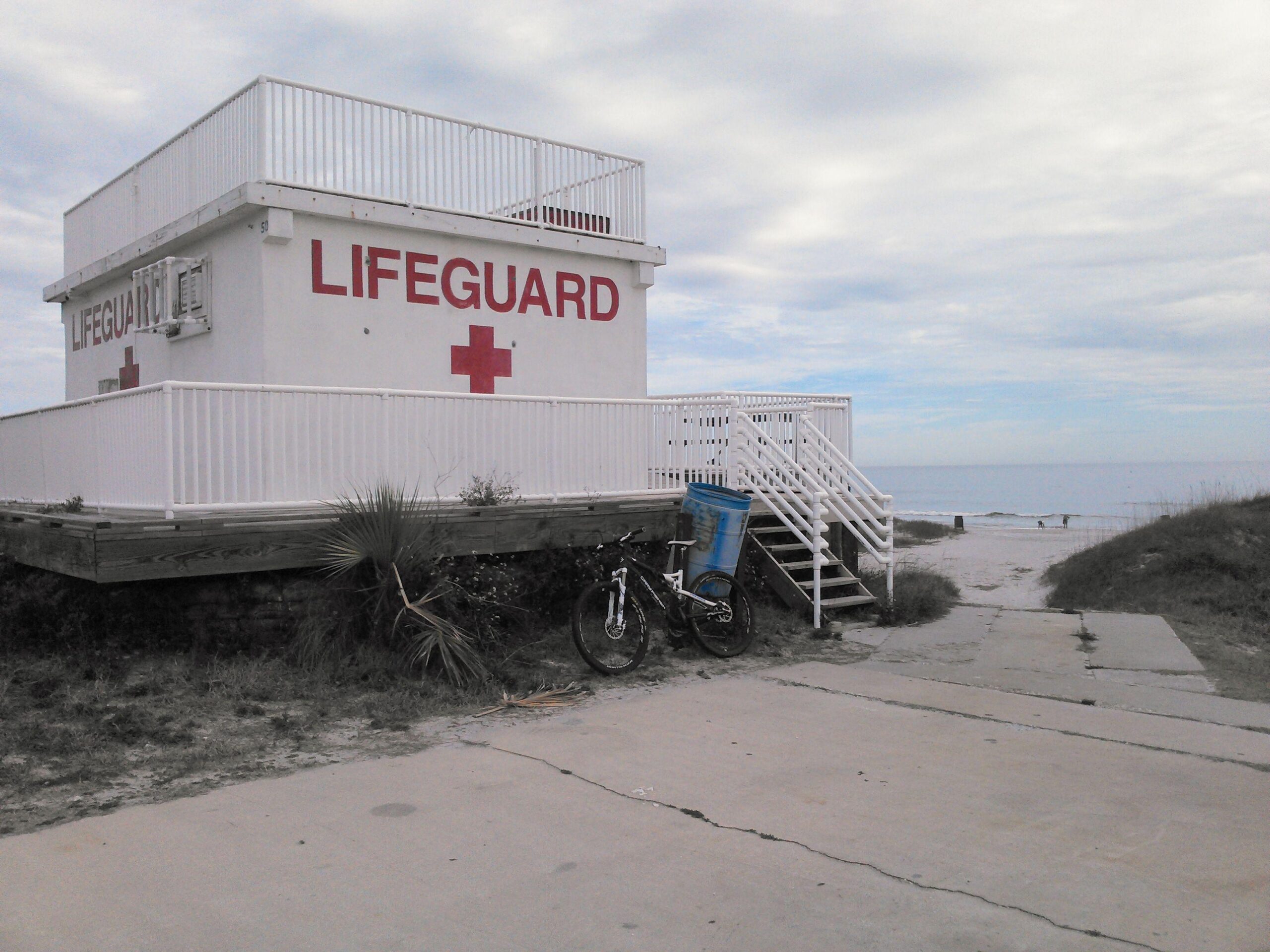 A lifeguard station painted in white with red lettering and a red cross, situated near the beach. A bicycle leans against the station, with a blue trash can nearby. The path leads to the sandy beach and ocean in the background, under a cloudy sky. Kathryn Abby Hanna Park mountain bike trail.