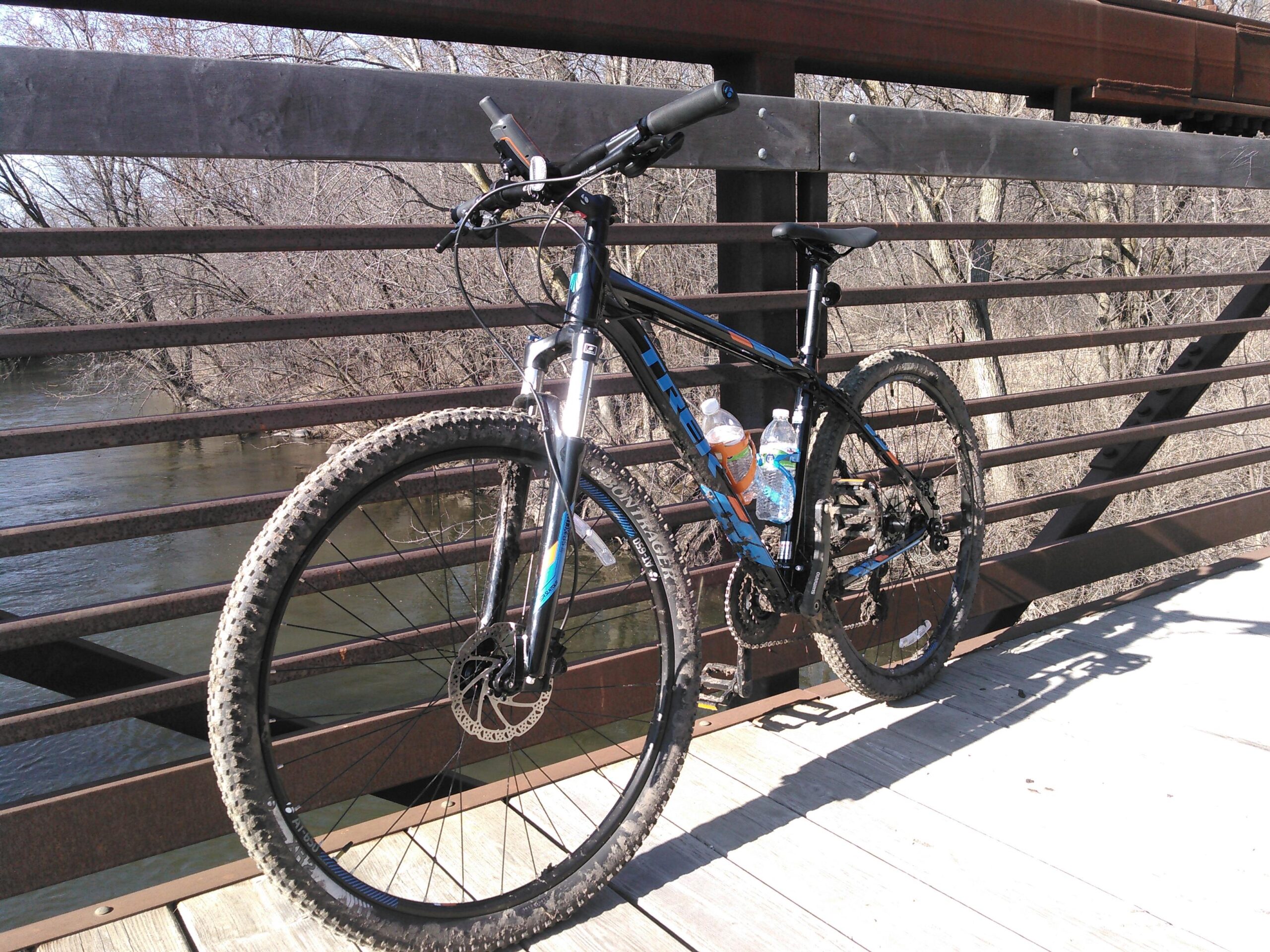 A mountain bike with a black frame and blue accents, resting on a wooden bridge. The bike has muddy tires and two water bottles attached to the frame. In the background, a calm river flows past bare trees, indicating a peaceful outdoor setting.