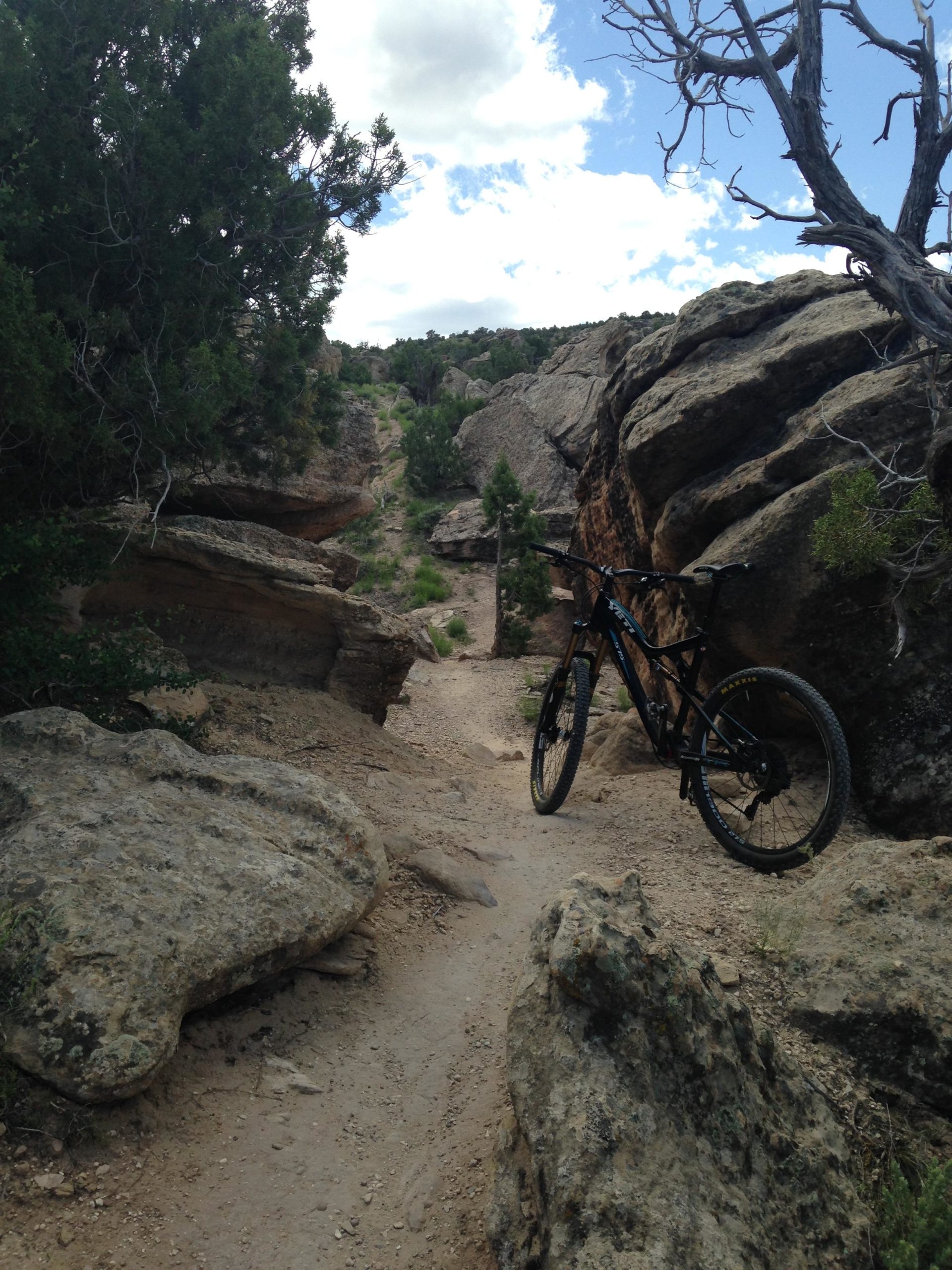 A black mountain bike parked on a dirt trail surrounded by large rocks and greenery. The path winds through a rocky landscape with a blue sky and scattered clouds above. Lunch Loops mountain bike trail.