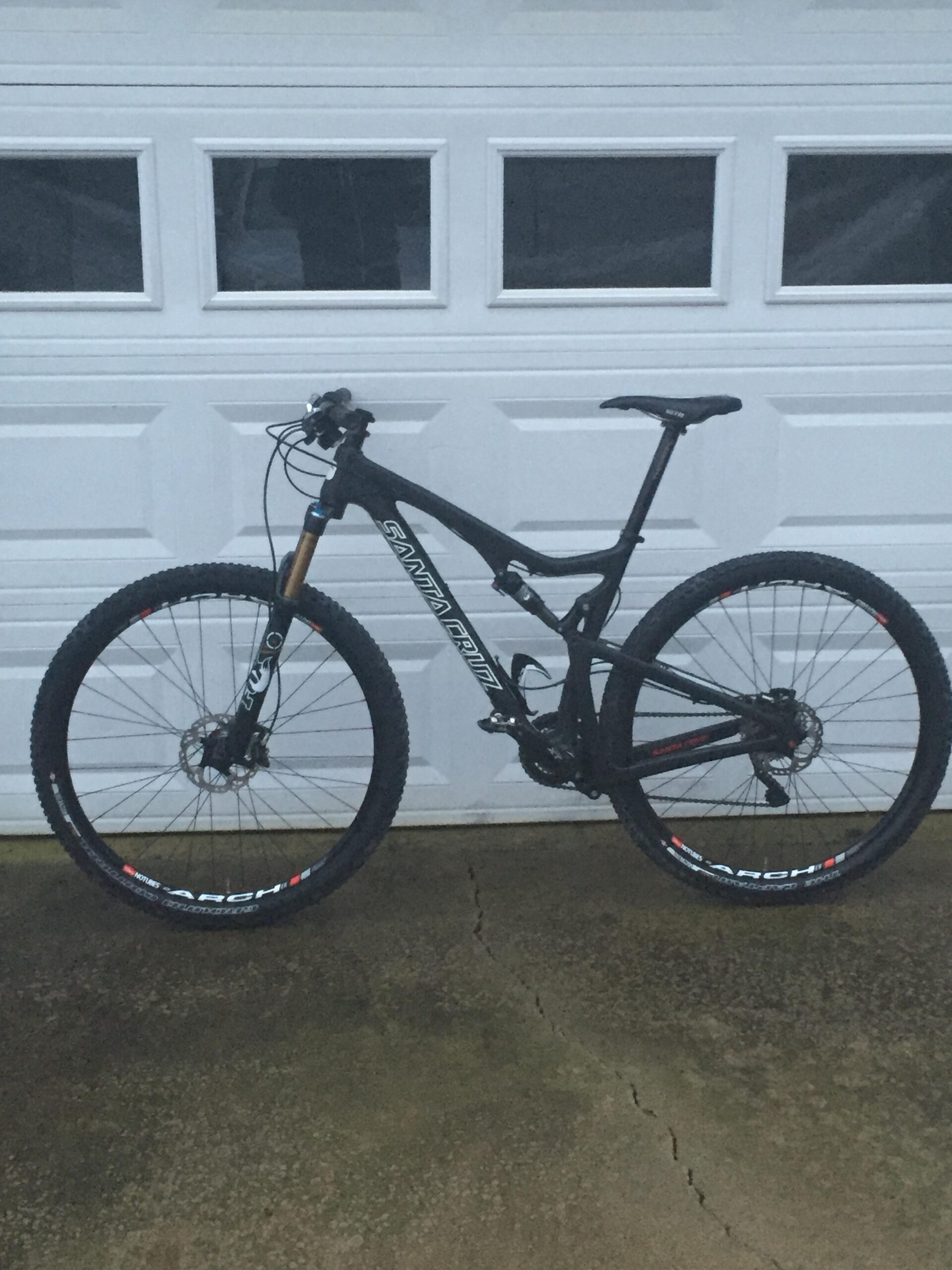 Santa Cruz Tallboy Carbon: A black mountain bike with thick tires and a sturdy frame, leaning against a garage door. The bike features front suspension and is set on a concrete surface, with visible cracks in the pavement.