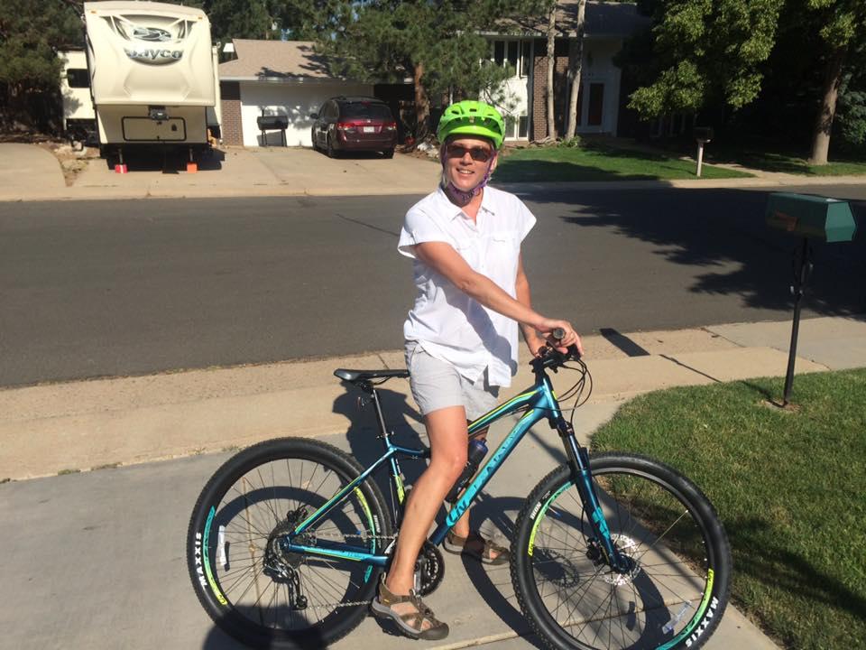 A person wearing a bright green helmet is smiling while sitting on a mountain bike on a sunny residential street. They are dressed in a white shirt and shorts, with grass and a mailbox visible in the foreground, and a house with a parked RV in the background.