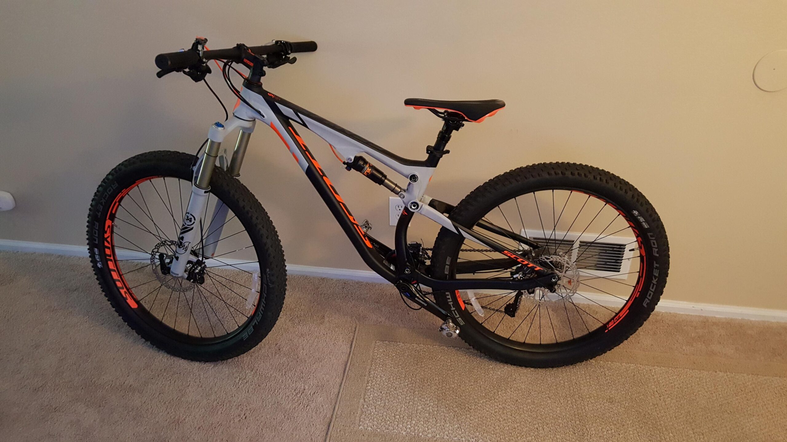 Scott Genius 720: A mountain bike with a sleek gray and black frame, featuring orange accents. The bike is shown upright on carpet, showcasing its large tires and front suspension forks. The seat is in a raised position, and the bike is positioned against a wall, highlighting its design and components.