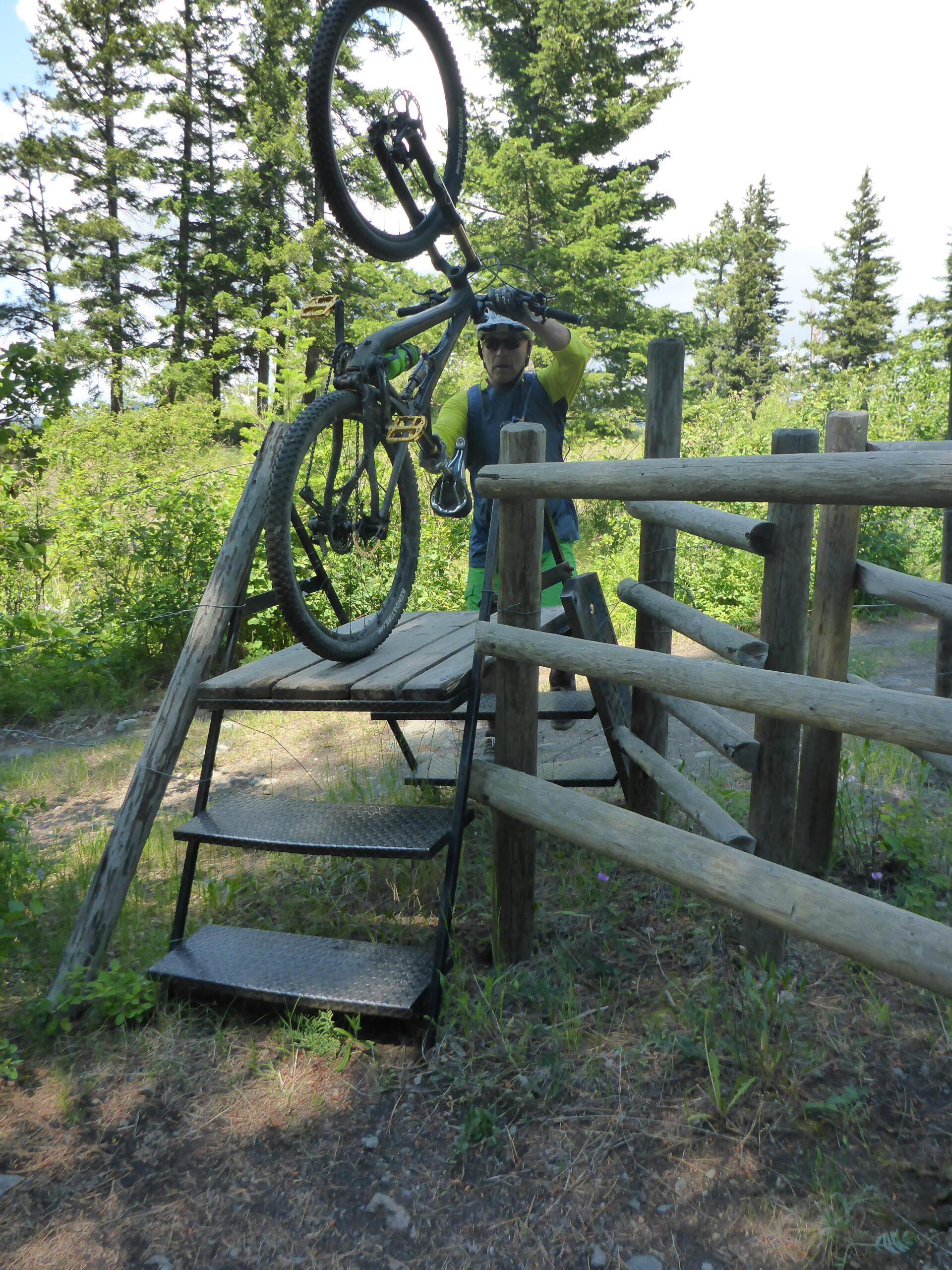 A person lifting a mountain bike over a wooden ramp with metal steps, surrounded by trees and greenery in a natural outdoor setting. The cyclist is wearing a helmet and sporty attire, preparing to navigate the ramp. Too Much Info Riding Area mountain bike trail.