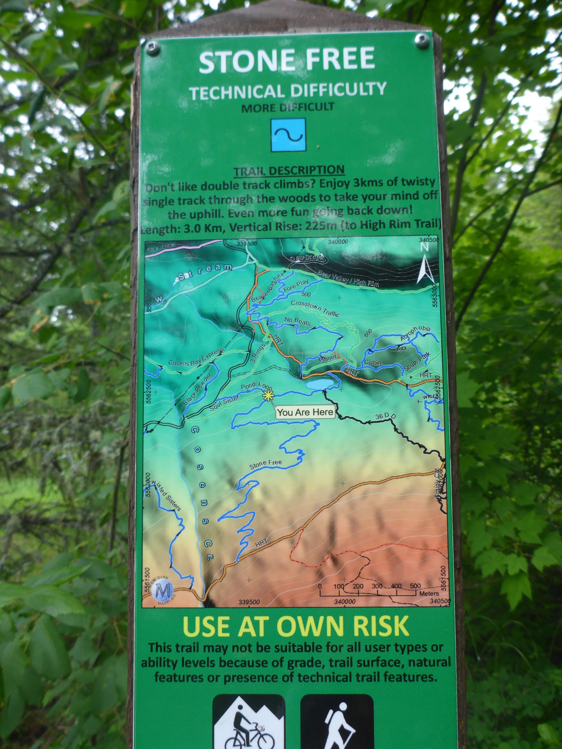 A sign displaying a trail map for the "Stone Free" hiking and biking trail, indicating it has a technical difficulty rating of "more difficult." The map shows various features, including terrain elevation, trail length of 3.0 km, and a vertical rise of 225 meters. Safety instructions advise users to "use at own risk," noting that the trail may not be suitable for all ability levels due to its grade and technical features. Cosens Creek Mountain Bike Area mountain bike trail.