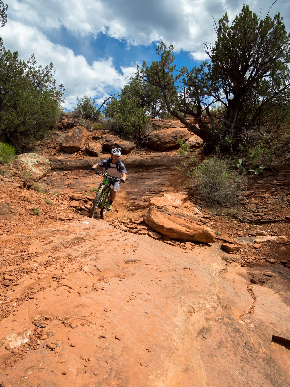 A mountain biker navigating a rocky trail in a desert landscape, surrounded by shrubs and trees under a partly cloudy sky. The terrain features reddish-brown rocks and a steep incline. Hiline mountain bike trail.