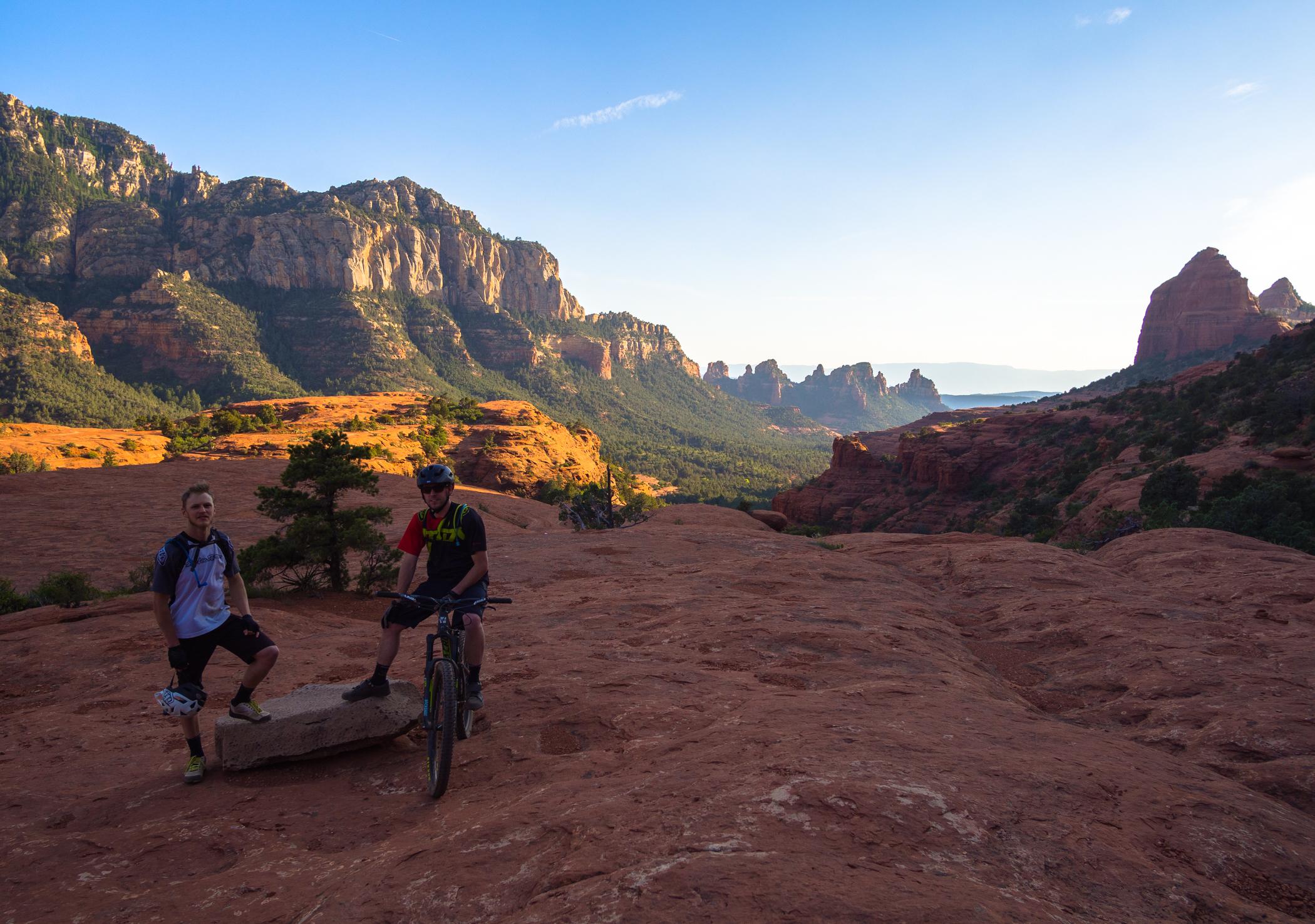 Two mountain bikers stand on a rocky outcrop in a desert landscape during sunset. One biker is posed next to a large stone, while the other is seated on his bike. The background features red rock formations and distant mountains under a blue sky with scattered clouds. Hangover mountain bike trail.