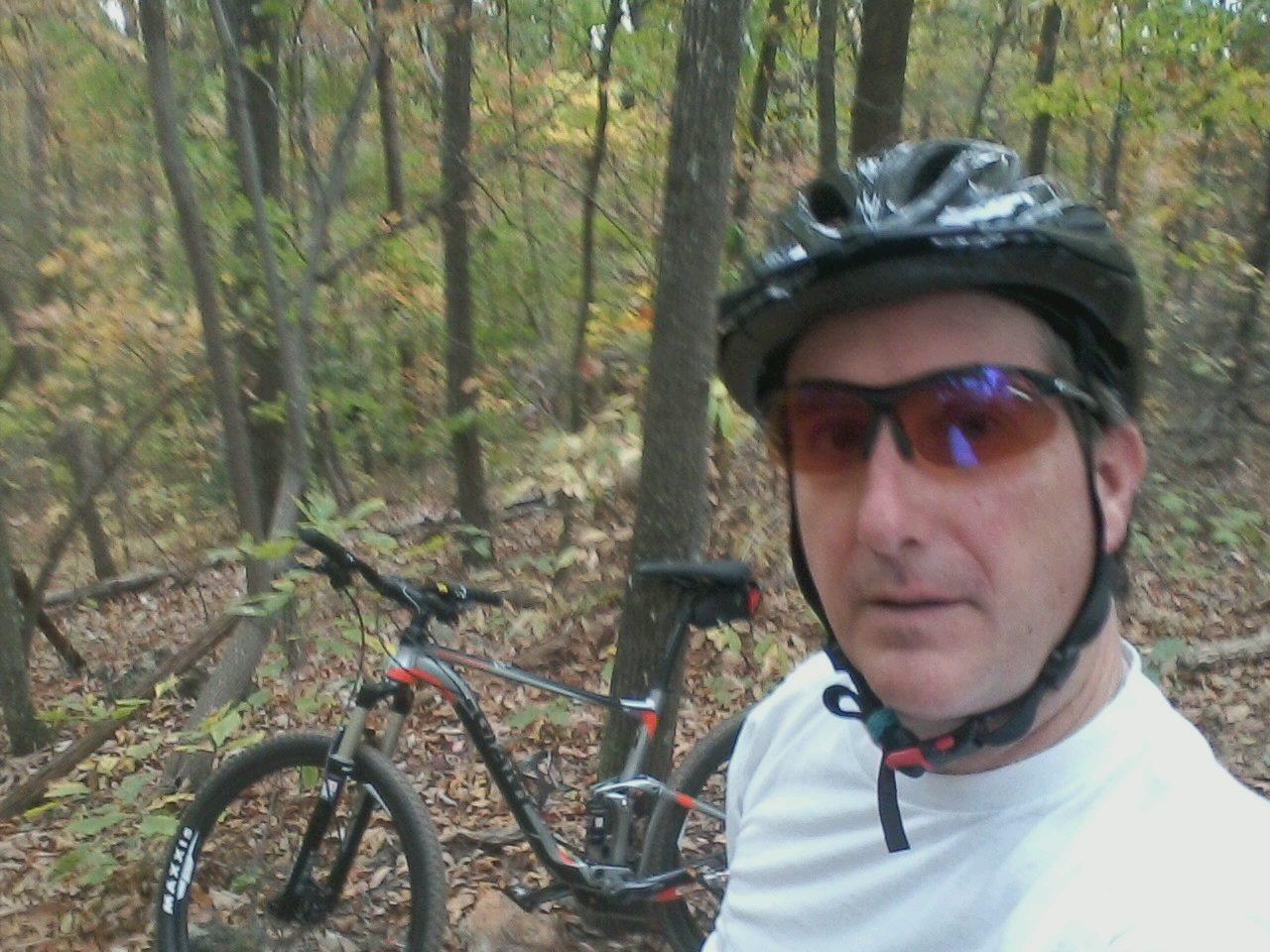 A person wearing a bike helmet and sunglasses poses for a selfie in a wooded area, with a mountain bike situated nearby. The background features trees and autumn foliage. Paris Mountain State Park mountain bike trail.