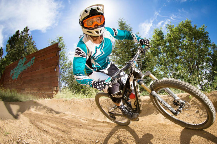 A mountain biker in a colorful jersey and protective gear leans into a turn on a dirt trail, with trees and a sign visible in the background. The sun shines brightly overhead, accentuating the action.