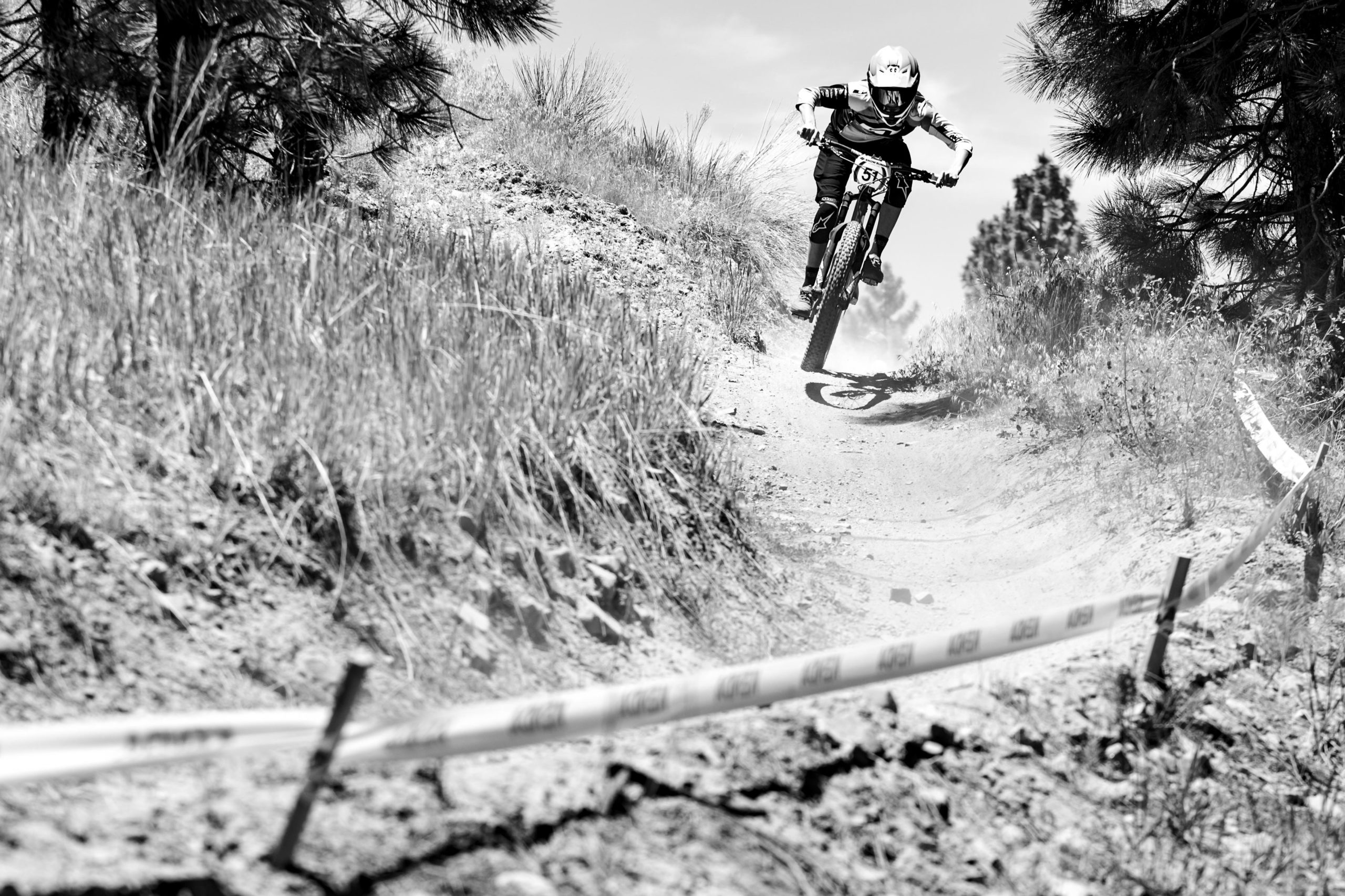 A mountain biker in mid-air jumps over a dirt trail surrounded by tall grass and pine trees, with a course boundary marked by tape visible on the ground. The image is in black and white, capturing the dynamic movement and excitement of the sport. Big Bear Mountain Resort mountain bike trail.
