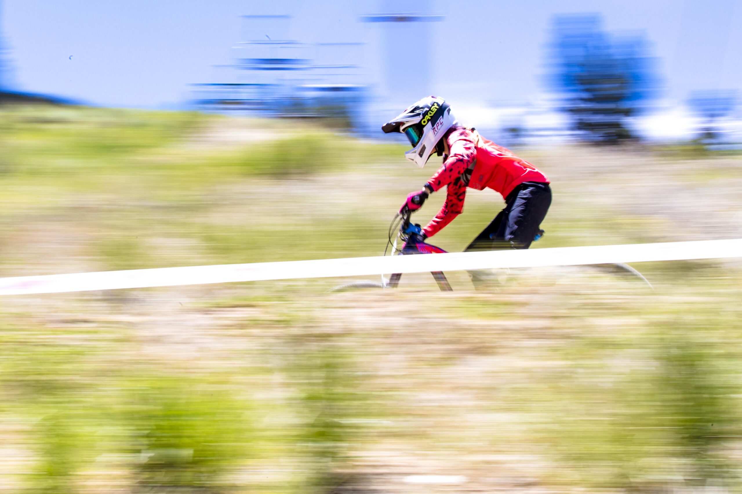 A mountain biker in a red jersey and helmet races downhill on a dirt trail, with motion blur emphasizing speed. The background features greenery and a clear blue sky. Big Bear Mountain Resort mountain bike trail.