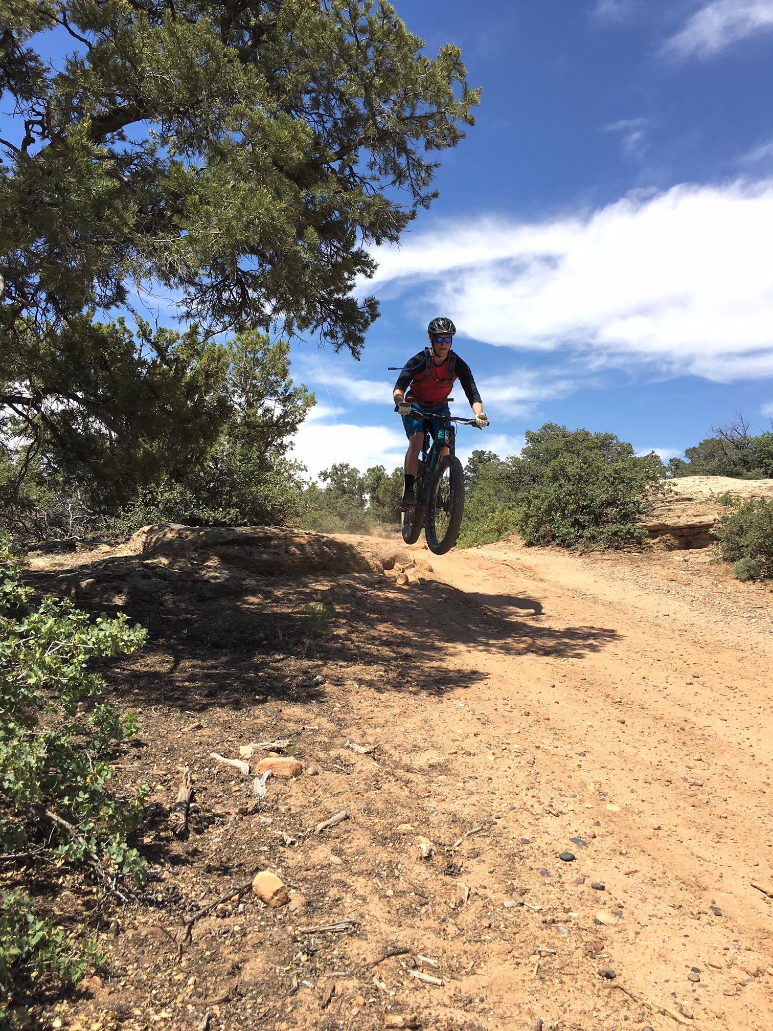 A mountain biker performing a jump over a rocky ledge on a dirt trail surrounded by shrubs and trees, with a blue sky and scattered clouds in the background. Gooseberry Mesa mountain bike trail.