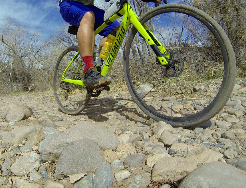 A close-up view of a mountain bike riding over rocky terrain, with a focus on the bike's front wheel and the rider's leg in motion. The bike features a bright yellow frame marked with "Specialized," and the rider is wearing blue shorts and red socks. The background includes scattered stones and dry vegetation under a blue sky. Road #45 mountain bike trail.