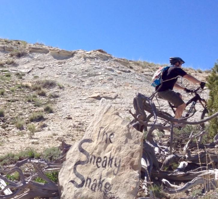 A mountain biker navigating a rocky trail with a sign labeled "The Sneaky Snake" in the foreground. The scene is set against a clear blue sky and rugged terrain. Tnt mountain bike trail.