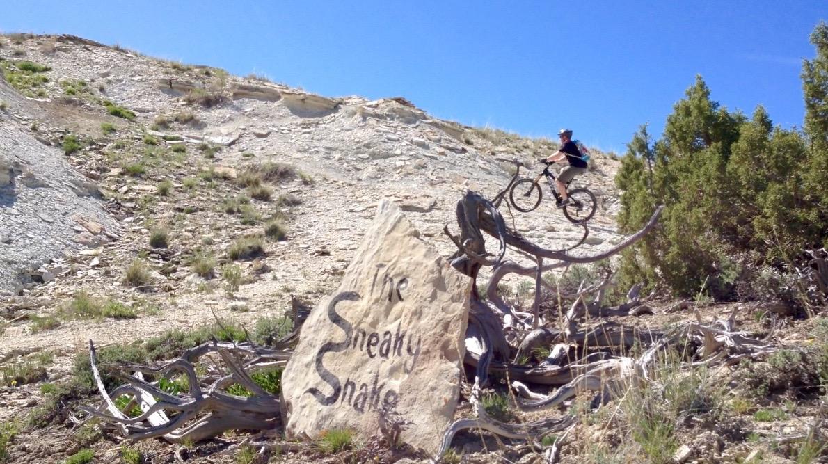 A mountain biker jumping over a rock with the inscription "The Sneaky Snake" on it, set against a steep, rocky hillside under a clear blue sky. Surrounding vegetation includes sparse grass and shrubbery. Tnt mountain bike trail.