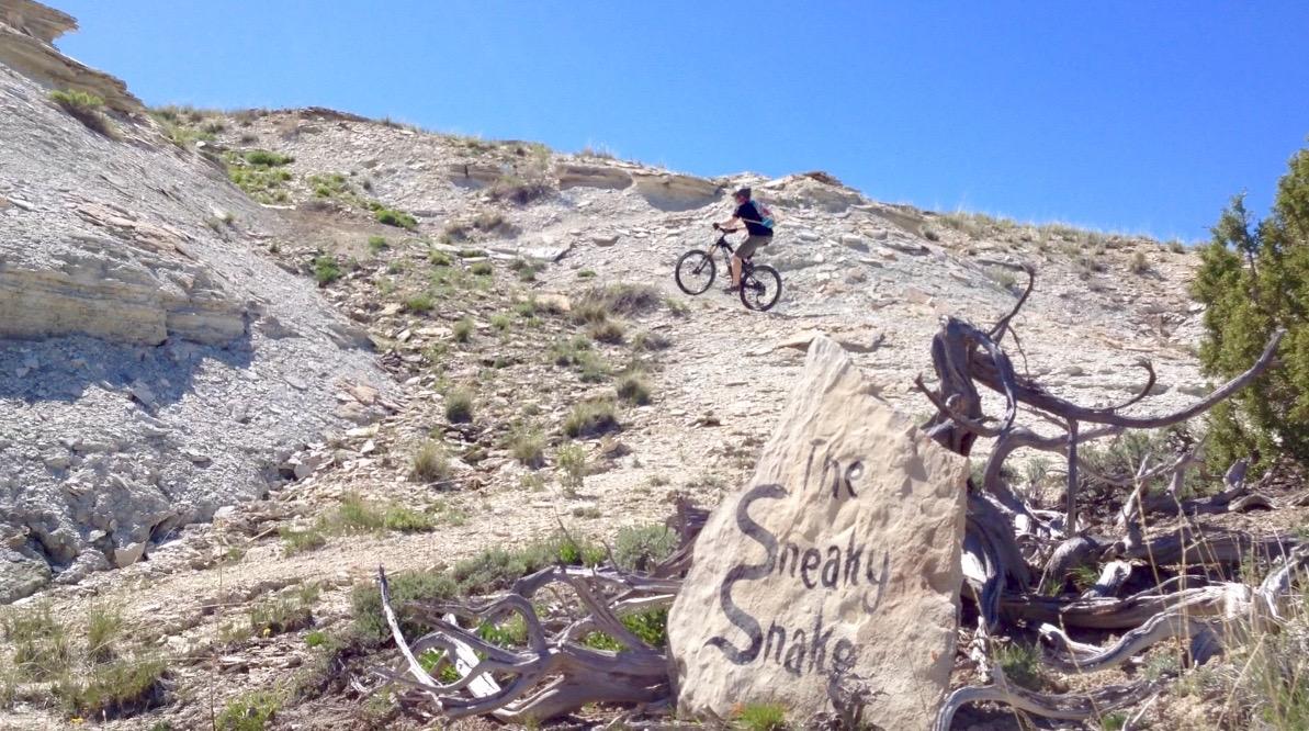 A mountain biker riding up a rocky slope with a clear blue sky in the background. In the foreground, a large stone marker reads "The Sneaky Snake," surrounded by sparse vegetation and fallen branches. Tnt mountain bike trail.