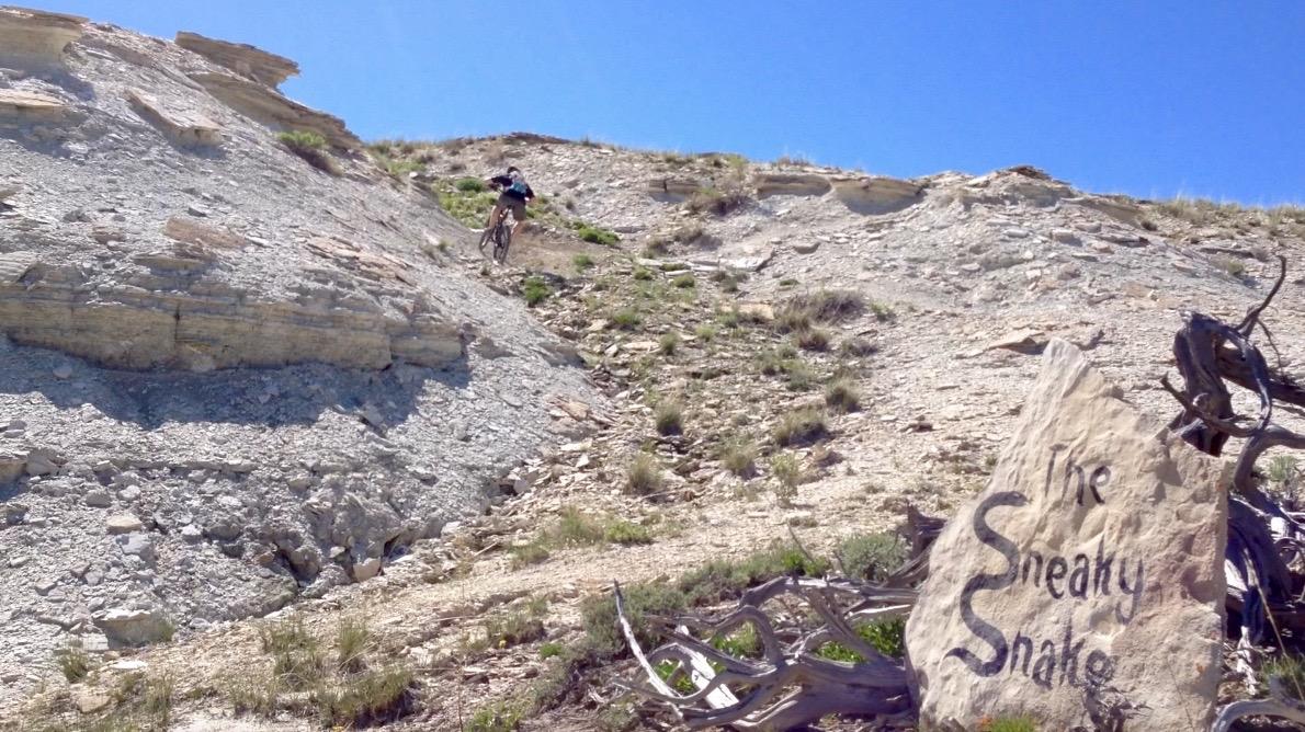 A mountain biker descending a rocky, sloped terrain with sparse vegetation, next to a large stone marked with the words "The Sneaky Snake" in a natural outdoor setting under a clear blue sky. Tnt mountain bike trail.