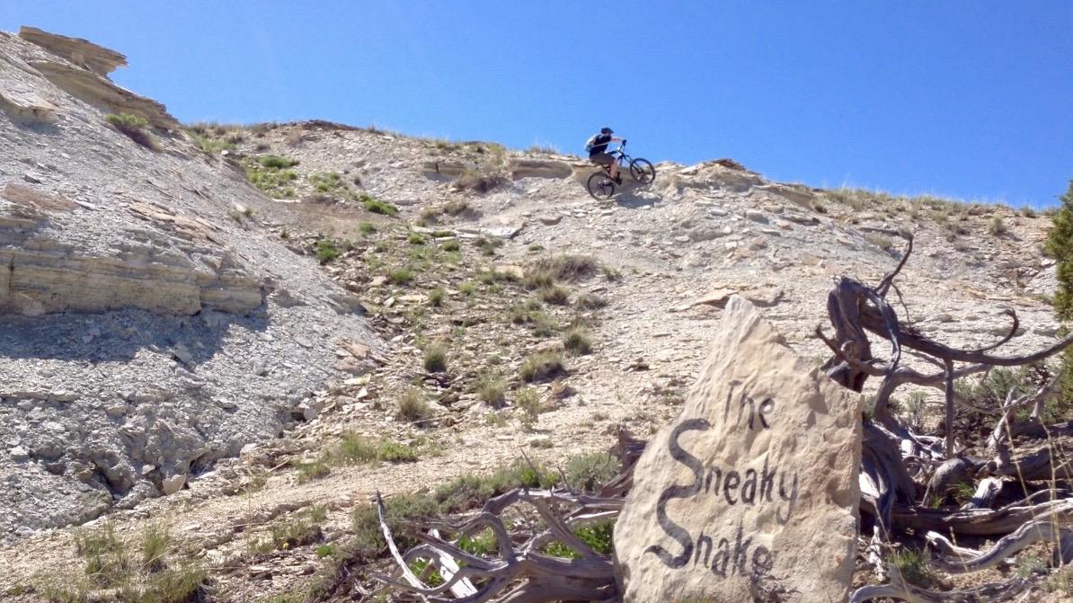 A mountain biker navigates a rocky terrain on a sunny day, with a sign reading "The Sneaky Snake" in the foreground. The landscape features gravel and sparse vegetation, indicating a rugged outdoor trail. Tnt mountain bike trail.