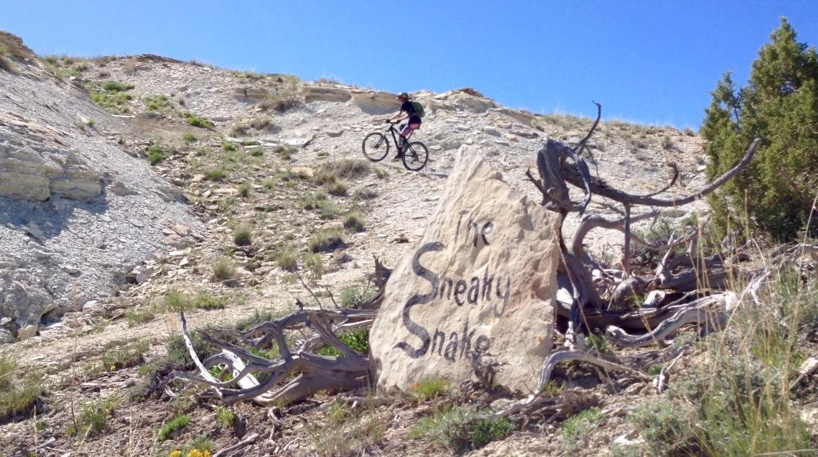 A mountain biker navigating a rocky trail, with a large stone marker reading "The Sneaky Snake" in the foreground. The scene is set against a clear blue sky, with sparse vegetation and steep terrain in the background. Tnt mountain bike trail.