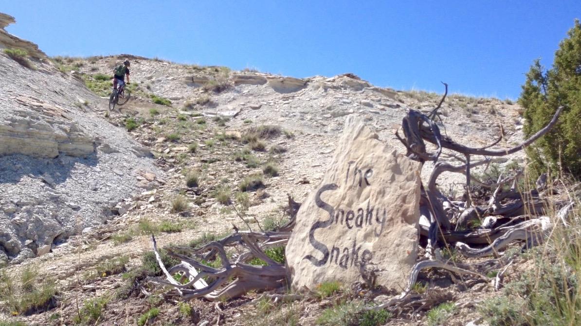 A mountain biker riding down a rocky slope next to a large stone with the inscription "The Sneaky Snake." The landscape features dry vegetation and rugged terrain under a clear blue sky. Tnt mountain bike trail.