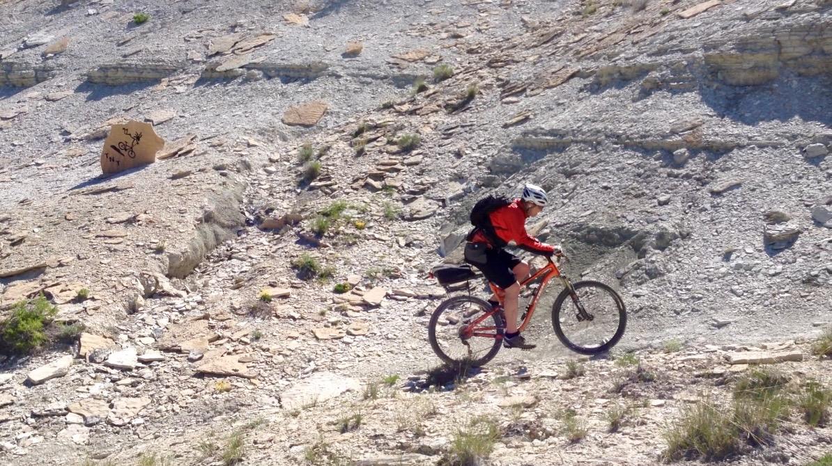 A mountain biker in a red jacket rides along a rocky trail, surrounded by uneven terrain and sparse vegetation. A large rock with markings is visible in the background, emphasizing the rugged landscape. Tnt mountain bike trail.