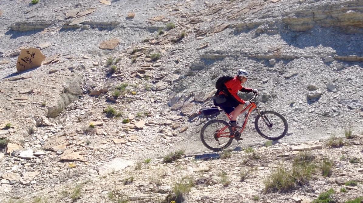 A mountain biker rides uphill on a rocky, gravel path surrounded by pebbles and sparse vegetation. The cyclist wears a helmet and a bright red jacket, and rides an orange mountain bike. A large, brown rock is visible in the background. Tnt mountain bike trail.