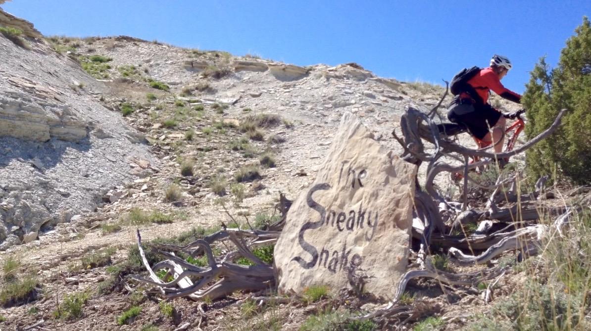 A cyclist riding on a rocky trail, near a large stone marker engraved with the words "The Sneaky Snake." The landscape features dry terrain with sparse vegetation and a clear blue sky in the background. Tnt mountain bike trail.