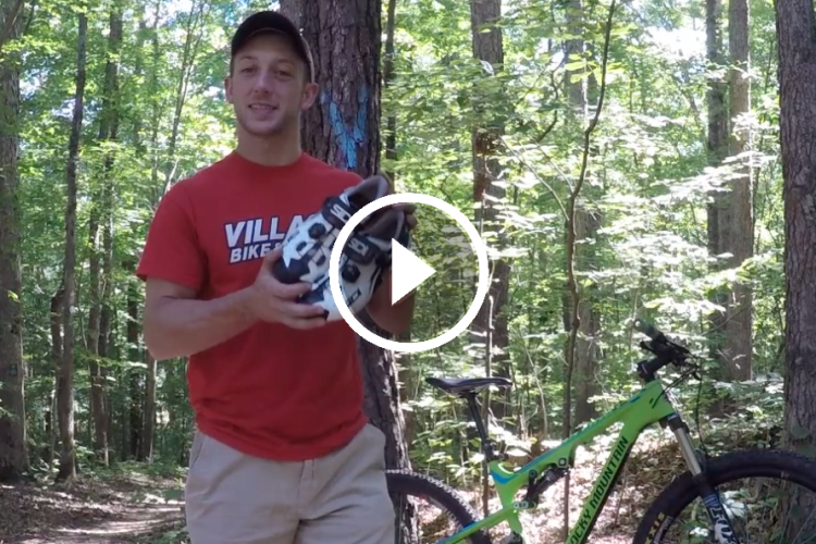 A person in a red t-shirt stands in a wooded area, holding a pair of cycling shoes. A green mountain bike is positioned next to them, and sunlight filters through the trees in the background. A play button icon indicates that this is a video.