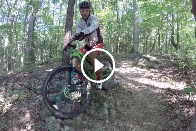 A mountain biker, wearing a helmet and protective gear, is positioned on a green bicycle on a rocky trail in a dense forest. The scene captures the natural surroundings with trees and sunlight filtering through the leaves. The biker appears focused, ready to navigate the challenging terrain ahead.