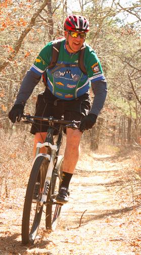 Salsa Horsethief: An older man riding a mountain bike along a wooded trail. He is wearing a colorful cycling jersey, shorts, gloves, and a red helmet, and is smiling as he enjoys the ride. The surrounding area features trees and dry foliage, indicating a sunny day.