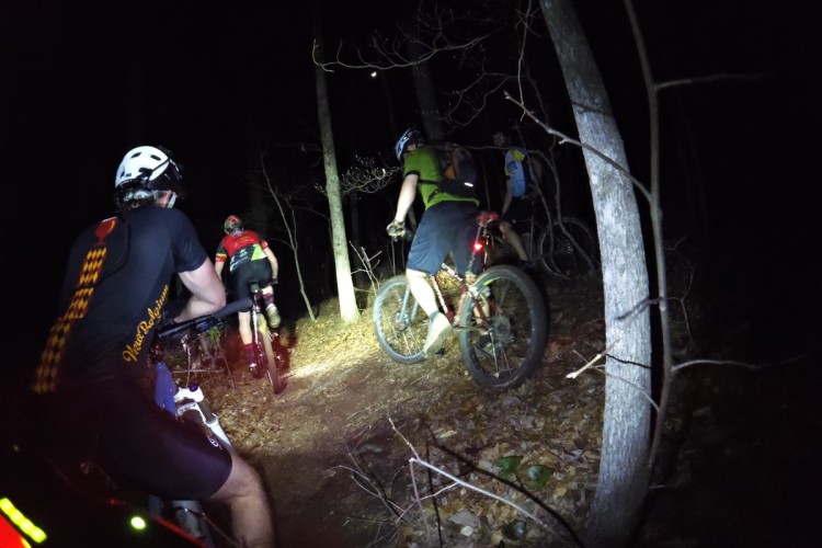 A group of mountain bikers riding along a narrow, leaf-covered trail in a dark forest, illuminated by bike headlights. The scene captures multiple riders wearing helmets and various cycling attire, with trees visible on either side of the path.