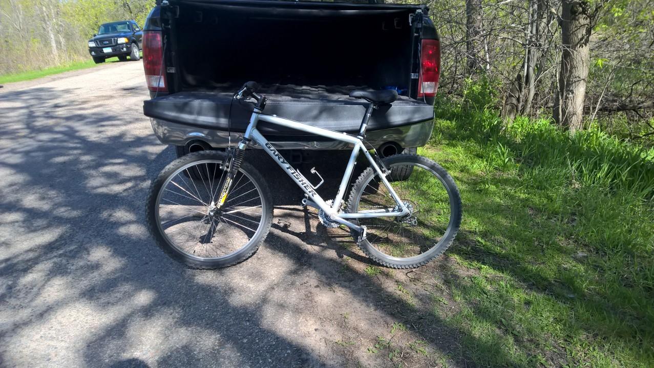Gary Fisher Tassajara: A white mountain bike with a front suspension is positioned next to the open tailgate of a black pickup truck. The bike is parked on a gravel road surrounded by green grass and trees, indicating a natural outdoor setting.