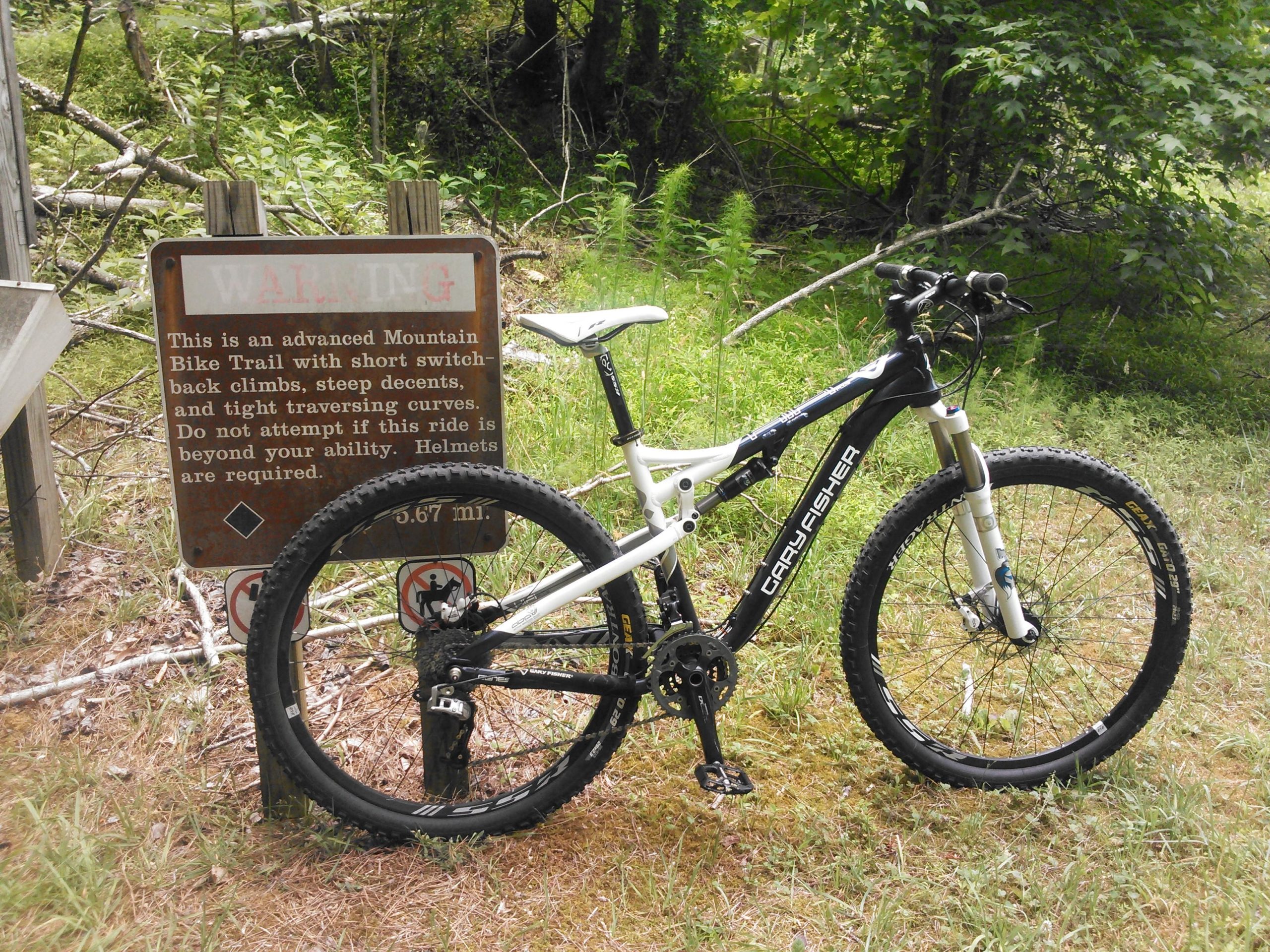 A mountain bike parked next to a warning sign indicating that the trail is advanced, featuring steep climbs, descents, and tight curves. The sign advises riders to use helmets and assess their skill level before attempting the trail. Lush greenery surrounds the area. York River State Park mountain bike trail.