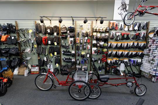 A red recumbent tricycle with a cargo basket in front is positioned in a bicycle shop. The background features shelves stocked with various cycling gear, including bags, helmets, and shoes, arranged neatly against the wall. The lighting enhances the vibrant colors of the accessories, creating a well-organized and inviting display for cycling enthusiasts.