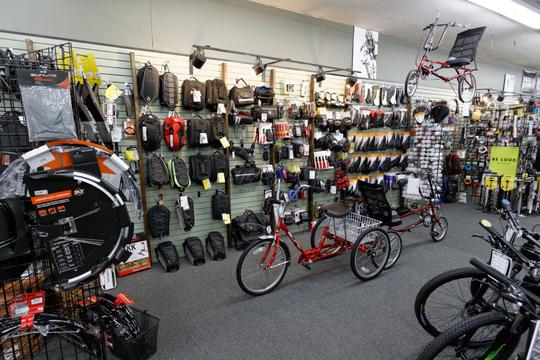 A retail store interior showcasing a variety of bicycles and cycling accessories. The image features a red three-wheeled bicycle in the center, surrounded by displays of backpacks, helmets, and other biking gear on the walls. Various bicycle tires and components are visible on the left side. The setting is well-lit and organized, highlighting the available products for cycling enthusiasts.