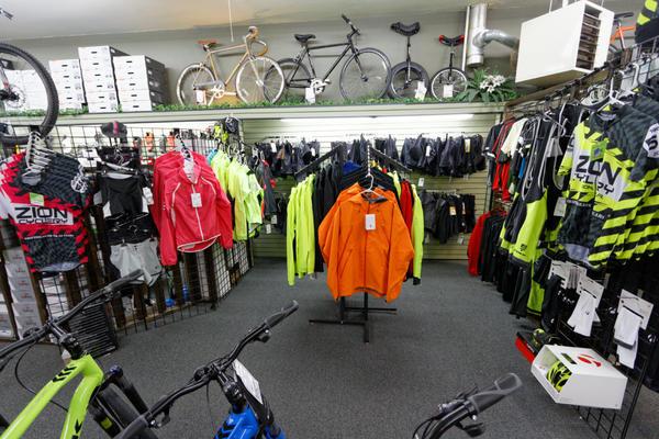 A well-organized bike shop interior showcasing a variety of cycling apparel and accessories. The image features brightly colored jackets, jerseys, and shorts displayed on racks, with bicycles hanging overhead. The shop is filled with items for cyclists, including helmets and other gear, all neatly arranged against a backdrop of grey flooring and walls.