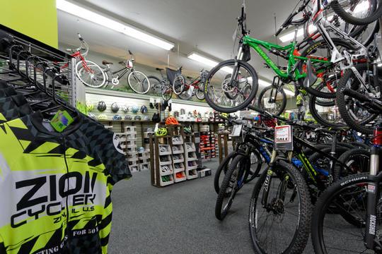 A vibrant cycling shop interior showcasing various bikes, including mountain bikes and children's bicycles, hanging from the ceiling and displayed on the floor. A rack of cycling apparel is in the foreground, featuring a jersey with "ZION CYCLERY" branding. The shop is well-lit and organized, with accessories, helmets, and a sale sign visible in the background.
