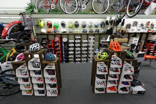 Interior of a bicycle shop featuring a variety of helmets displayed on wooden shelving units. The walls are lined with more helmets in different colors, and there are biking accessories and shoes arranged on shelves. Two rows of stacked boxes are seen in the foreground, showcasing additional helmet options. Bicycles hang from the ceiling in the background.