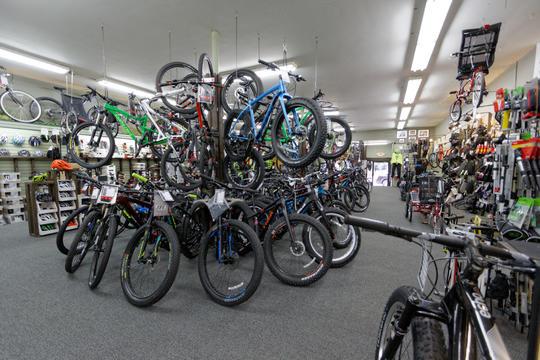 An indoor bicycle shop filled with various models of bikes hanging from the ceiling and displayed on the floor. The space features bright lighting, carpeted flooring, and a wall lined with cycling accessories. Several customers are visible browsing the merchandise.