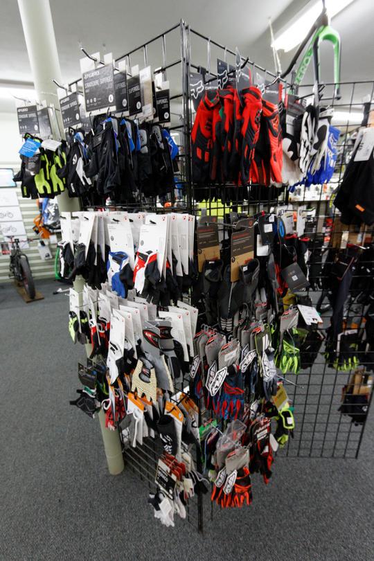 A display rack featuring various styles and colors of gloves, including biking and sports gloves, organized by category. The rack is filled with gloves in a range of colors, including blue, red, black, and green, with packaging labels visible. The background shows part of a retail store, with additional sporting goods in view.