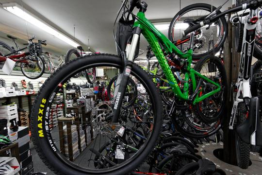A crowded bicycle shop displaying a variety of bicycles, including a prominent green mountain bike with thick tires. The background features numerous other bikes hanging from displays, and shelves stocked with bike accessories and gear.