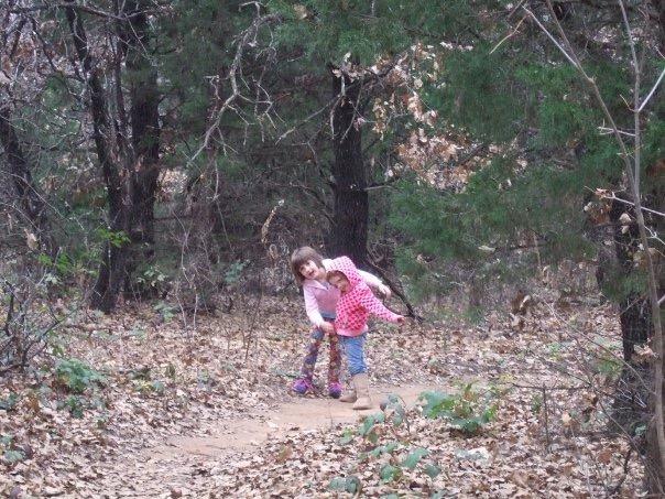 Two children playing together on a dirt path through a wooded area, surrounded by trees and fallen leaves. One child is wearing a pink sweater and blue jeans, while the other is in a pink patterned shirt and purple leggings, both looking joyful. Arcadia Laketrails mountain bike trail.