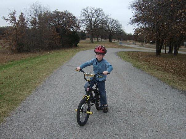 A young boy wearing a red helmet is riding a black bicycle on a gravel path surrounded by trees. He is facing the camera and wearing a blue long-sleeve shirt and jeans, with a grassy area on either side of the path. The scene is set on a cloudy day. Arcadia Laketrails mountain bike trail.
