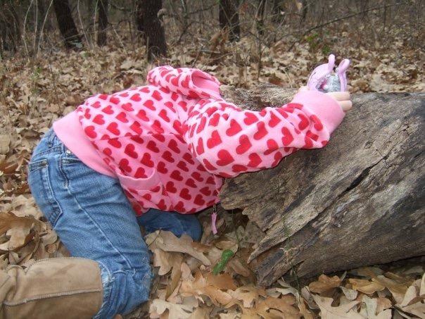A child in a pink hoodie with a heart pattern is playfully leaning over a log in a wooded area covered with fallen leaves. Arcadia Laketrails mountain bike trail.