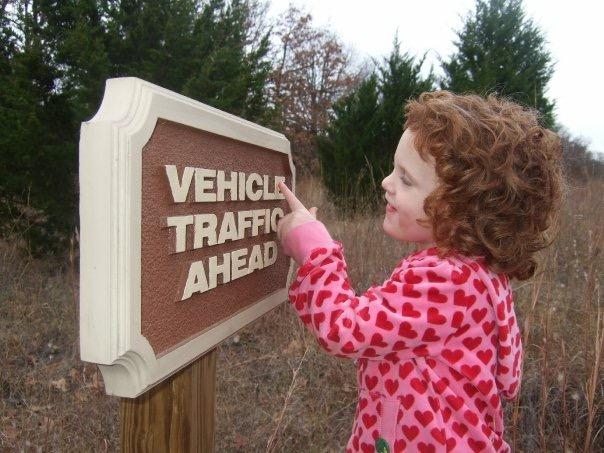 A young child with curly red hair, wearing a pink sweatshirt with heart patterns, points at a road sign that reads "VEHICLE TRAFFIC AHEAD." The sign is mounted on a wooden post, surrounded by a natural landscape with evergreen trees in the background. Arcadia Laketrails mountain bike trail.