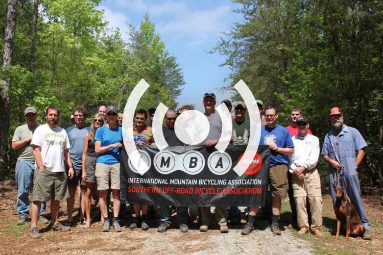 A group of individuals poses outdoors, standing together in a forested area. They are holding a banner for the International Mountain Bicycling Association (IMBA) and the Southern Off-Road Bicycle Association (SORBA). The group consists of men and women of various ages, some wearing hats and casual clothing, with a dog sitting by one of the members. The background features green trees and a gravel path.