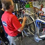 A person in a red shirt is working on a bicycle in a workshop, surrounded by various bike parts and tools. Other individuals can be seen in the background.